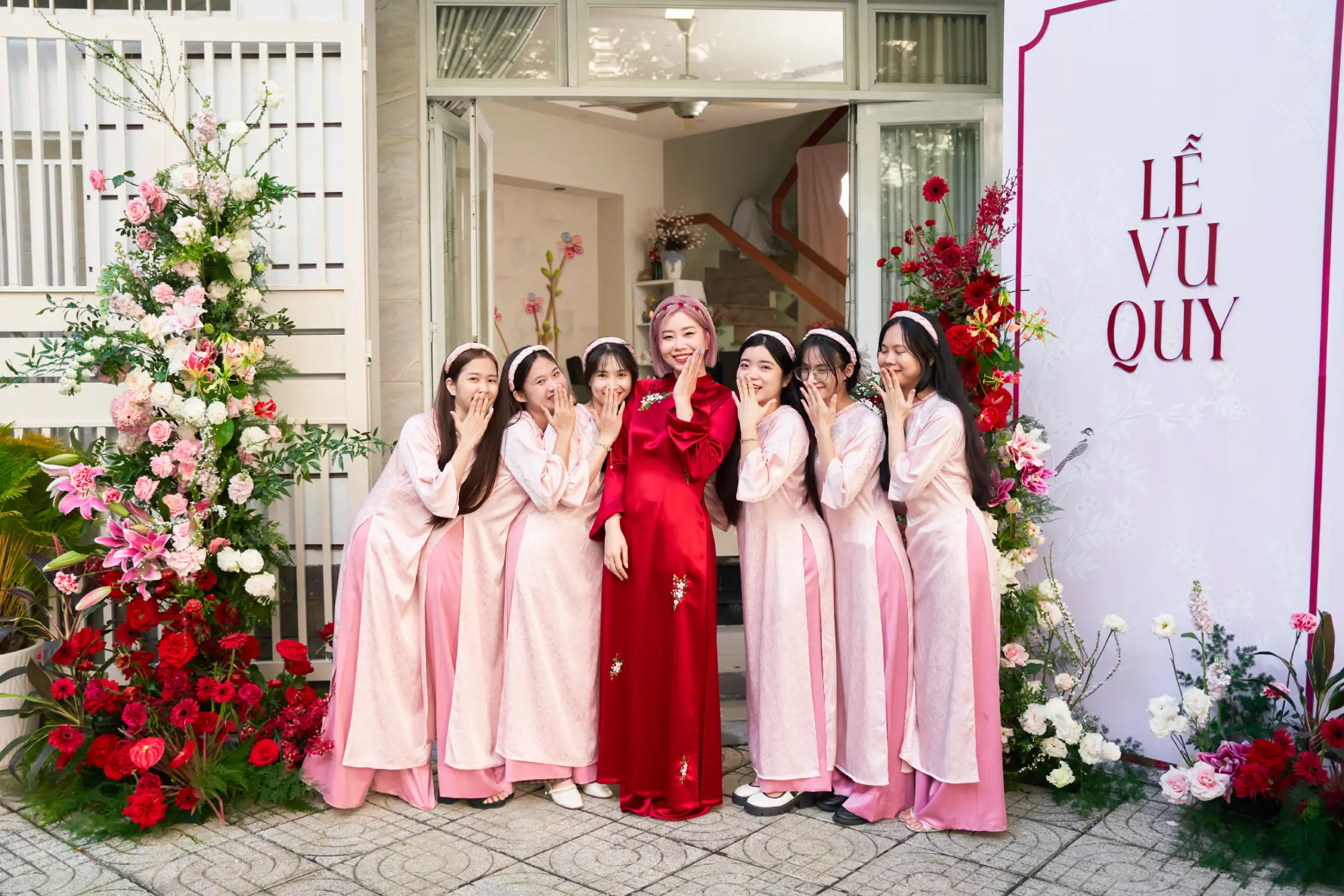 Cô dâu trong áo dài đỏ rực rỡ đứng cùng dàn phù dâu mặc áo dài hồng phấn trước lễ vu quy, khoảnh khắc vui tươi và tràn ngập năng lượng.