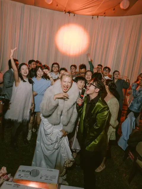 The bride laughing and singing along with a guest holding a microphone as friends gather around during the reception.