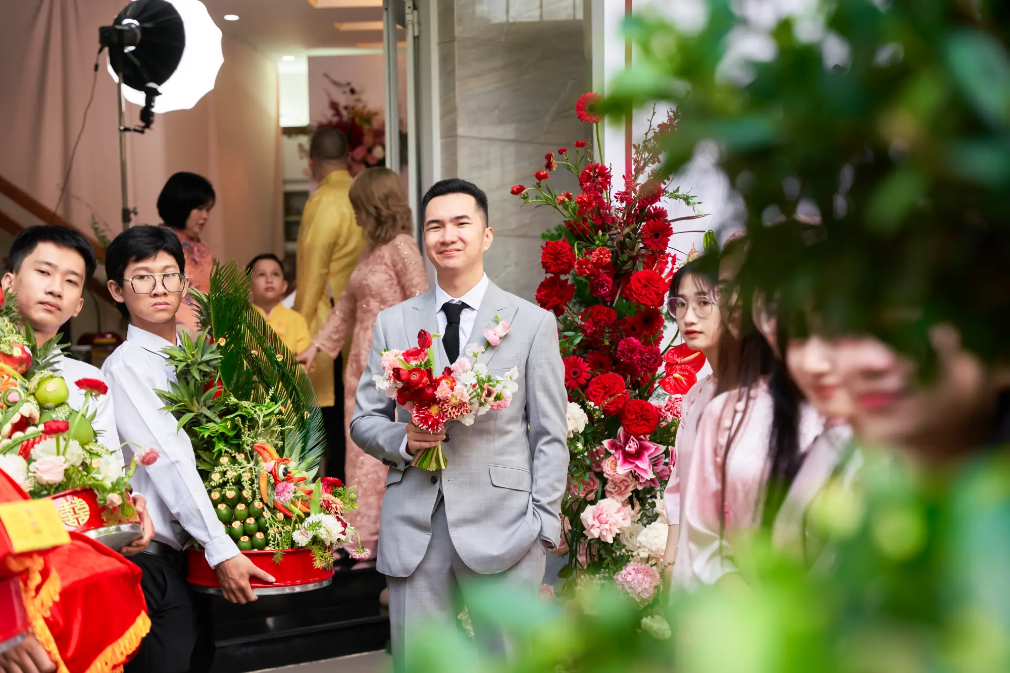 Chú rể trong bộ vest xám cầm bó hoa cưới đỏ, cùng đội bưng quả mang tráp trầu cau và sính lễ đến nhà gái trong không khí lễ cưới rộn ràng.