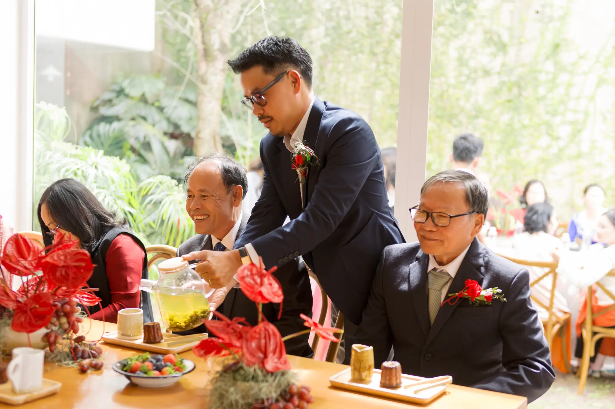 Groom Duy serving tea to family elders, surrounded by red floral decorations in a warm and intimate setting.