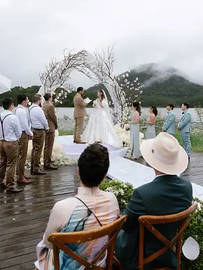 Couple exchanging vows on a wooden deck by Tuyen Lam Lake in Da Lat, framed by a white arch adorned with branches and blossoms, with misty mountains in the background and bridal party standing alongside