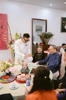 Chú rể rót trà mời cha mẹ trong nghi thức lễ gia tiên tại nhà gái, cả gia đình quây quần bên bàn tròn với hoa tươi và trà bánh.