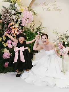 Bride holding hands with a young flower girl while posing in front of a colorful floral arrangement.