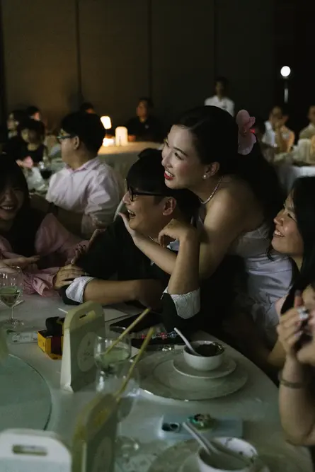 Bride leaning over a friend’s shoulder and laughing with guests at a reception table.