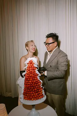 Cặp đôi cười rạng rỡ bên bánh cưới phủ dâu tây trong không gian tiệc tối ấm áp.