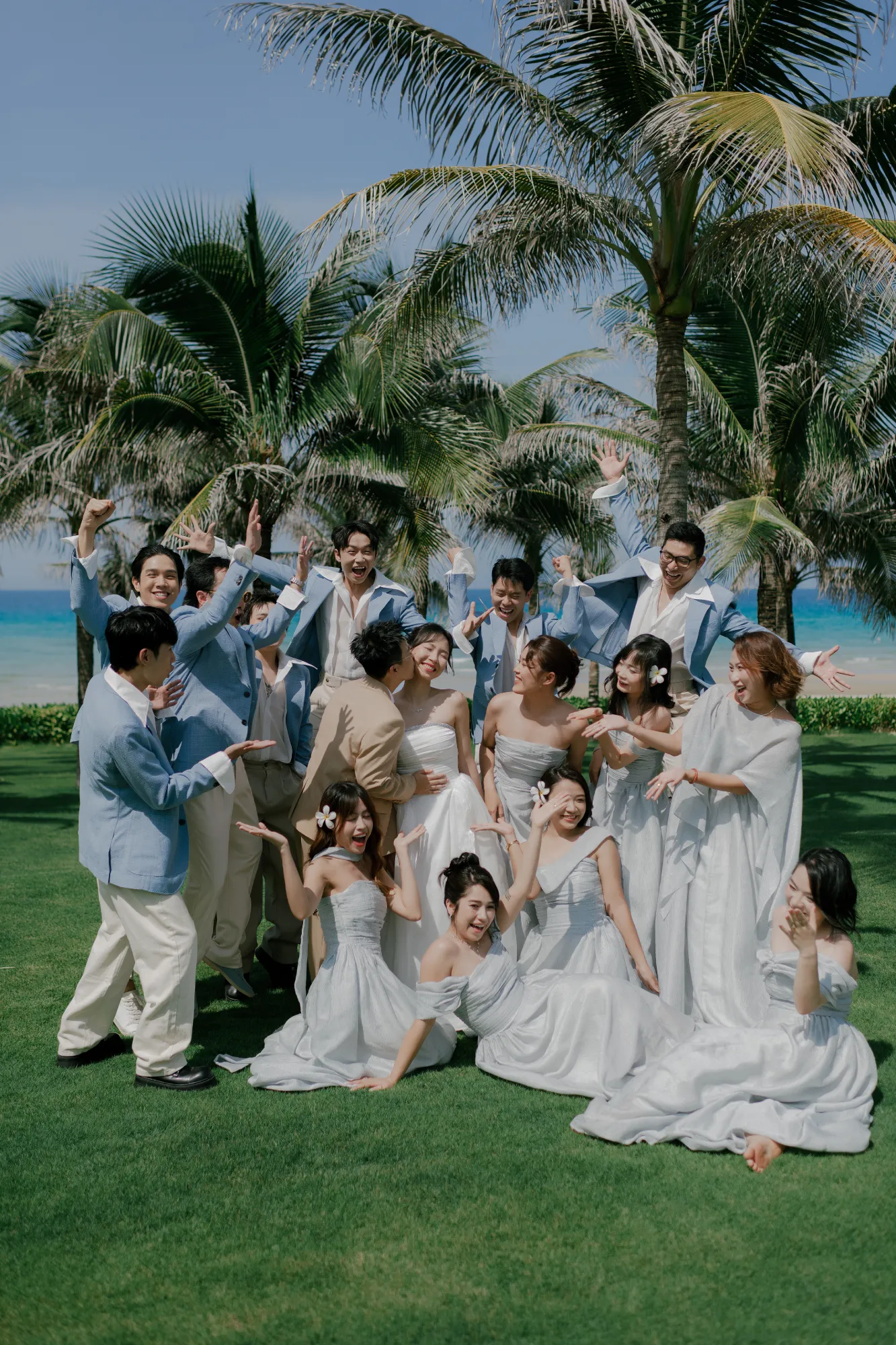 Cô dâu chú rể cùng dàn phù dâu phù rể tạo dáng rộn ràng trên bãi cỏ xanh bên bờ biển, tràn đầy năng lượng và tiếng cười hạnh phúc.
