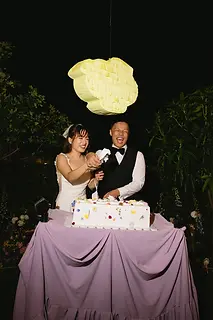 A joyful cake cutting moment with laughter during an outdoor evening wedding celebration.