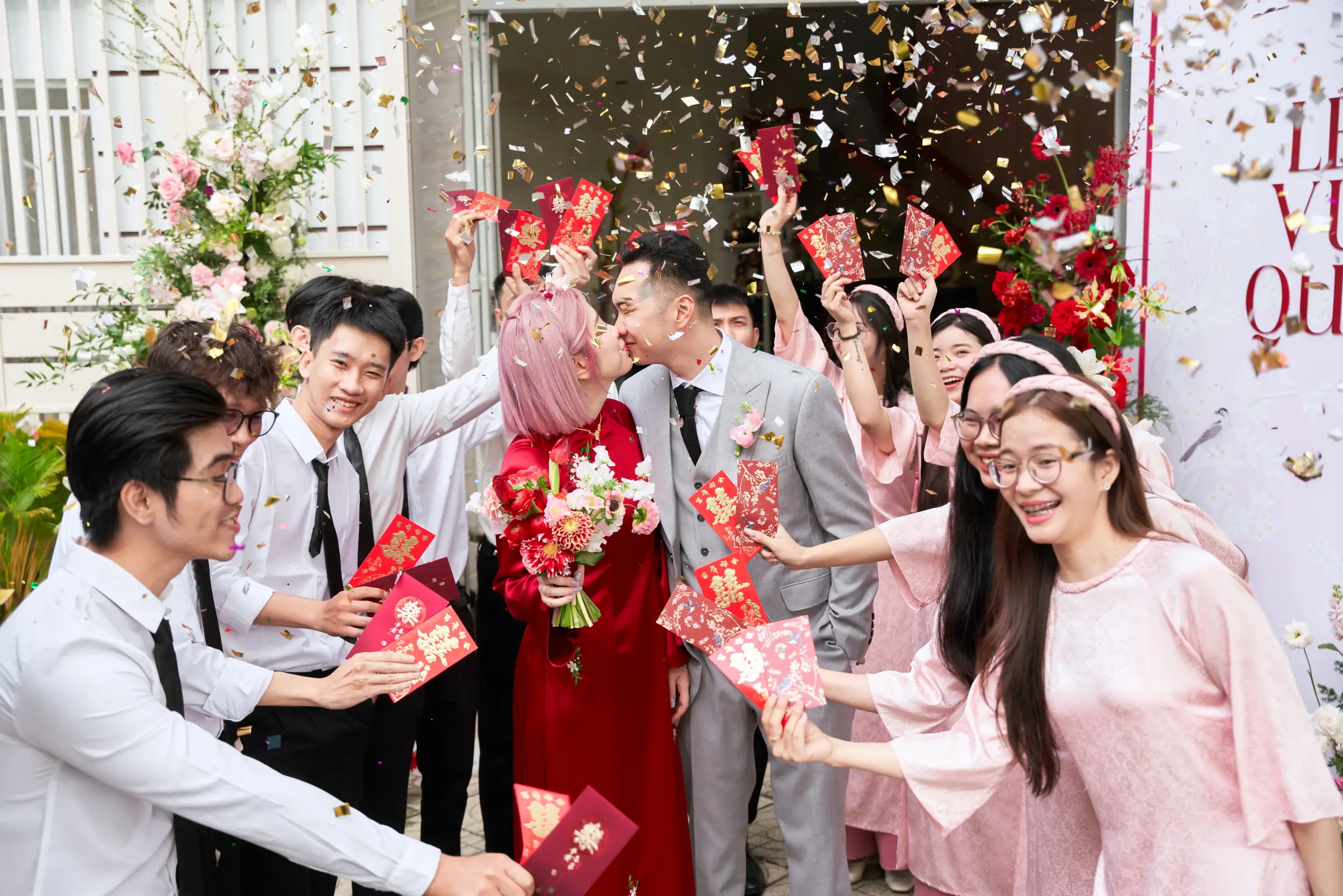 Bride and groom exchanging a kiss as friends shower them with confetti and red envelopes, celebrating the Vietnamese lucky money tradition (Lì Xì Trả Duyên).
