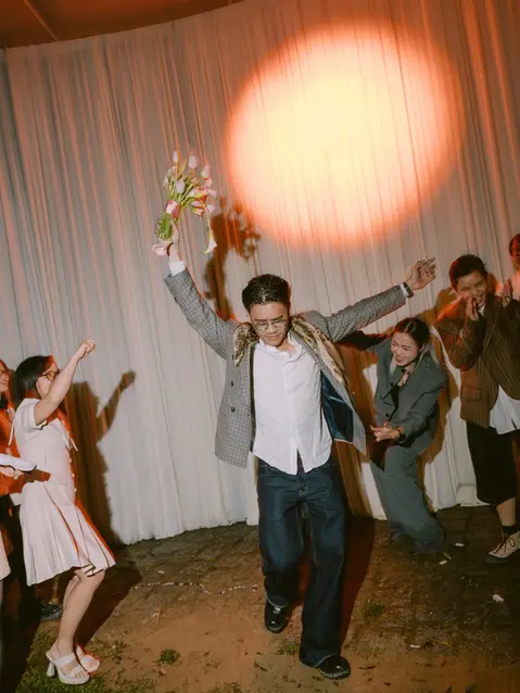 Khách mời cầm bó hoa nhảy giữa sàn tiệc, mọi người xung quanh cổ vũ trong không khí vui nhộn và sôi động.
