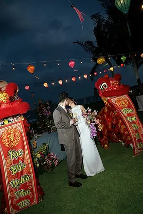 Newlyweds toasting and kissing between red lion dance banners of blessings at their evening beachfront reception