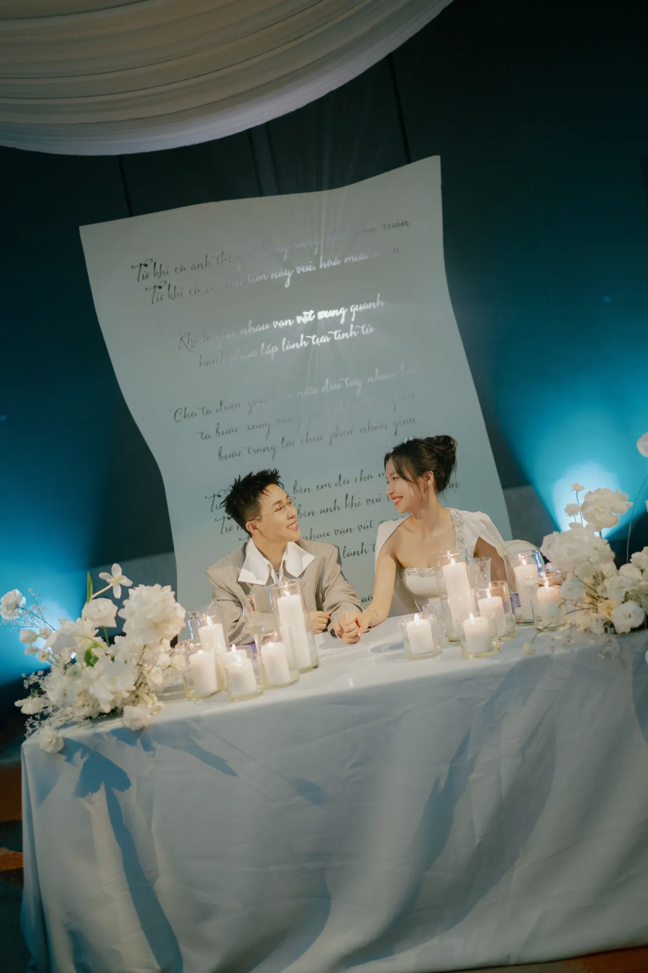 Cô dâu và chú rể bên bàn tiệc ngập ánh nến, ánh mắt trao nhau đầy tình cảm trong không gian ấm cúng.