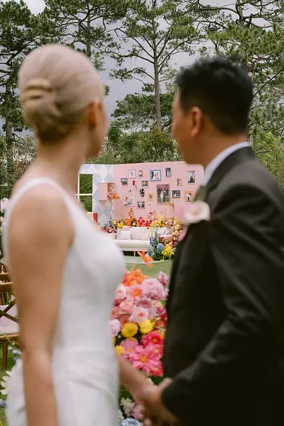 Cặp đôi nắm tay nhìn về sân khấu lễ cưới ngoài trời được trang trí bằng hoa nhiều màu sắc.