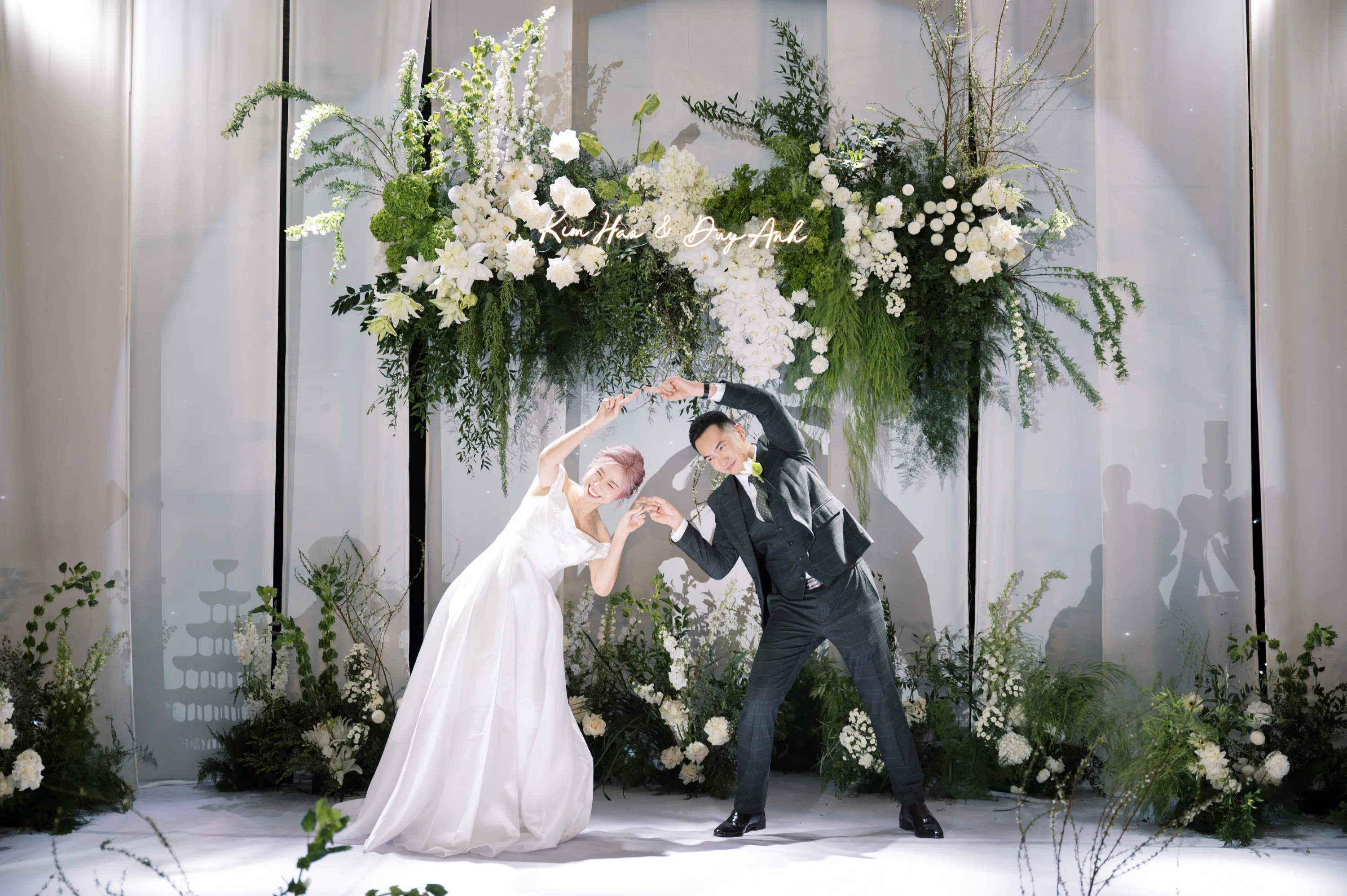 Cặp đôi Kim Hảo & Duy Anh tạo dáng vui nhộn trên sân khấu lễ cưới, giữa không gian hoa trắng tươi tốt và ánh sáng lung linh.