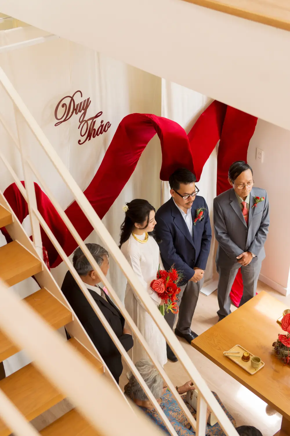 Cô dâu chú rể trong lễ trà rượu gia tiên tại nhà, không gian được trang trí tối giản với dải lụa đỏ mềm mại và bảng tên.