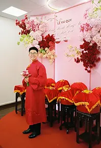 Groom in red áo dài holding calla lilies beside red-covered traditional gift trays at a Vietnamese wedding ceremony