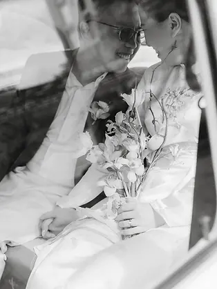 Black and white portrait of a bride and groom sitting closely together inside a vintage car.