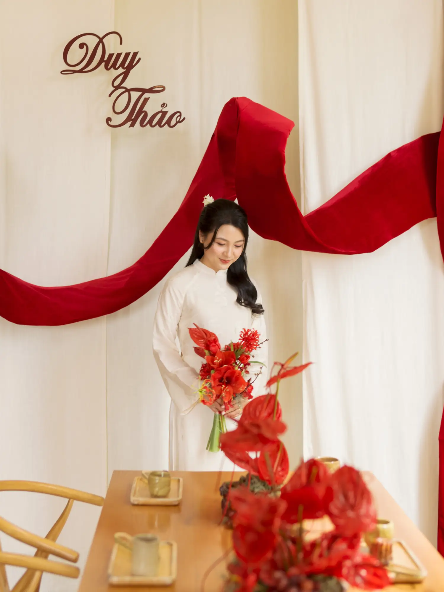 Cô dâu trong áo dài trắng truyền thống cầm bó hoa đỏ, đứng trước phông nền trang trí với dải lụa đỏ mềm mại, toát lên vẻ thanh lịch và nhẹ nhàng.