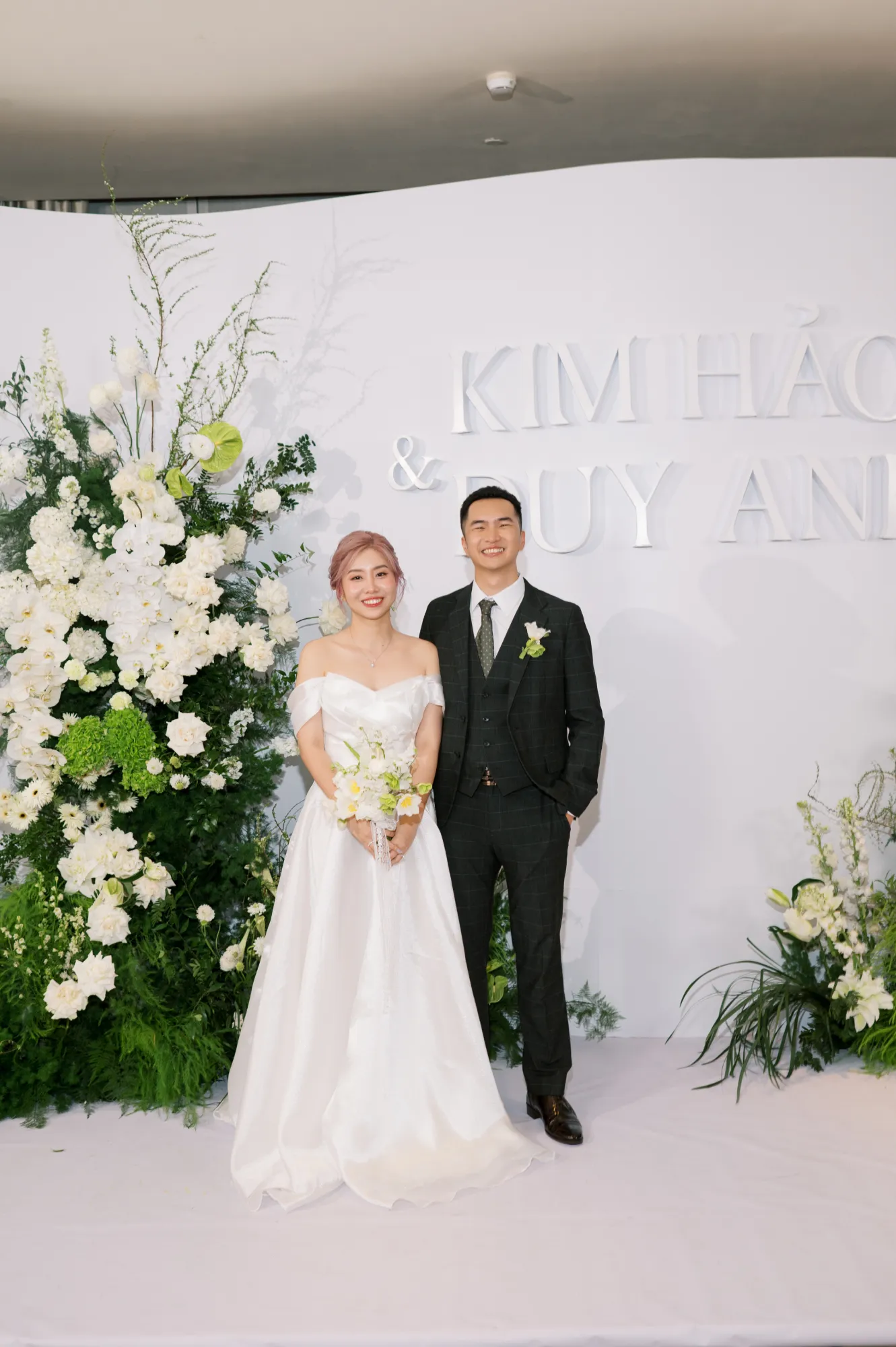 Bride and groom Kim Hao & Duy Anh posing in front of a modern white floral wedding backdrop.