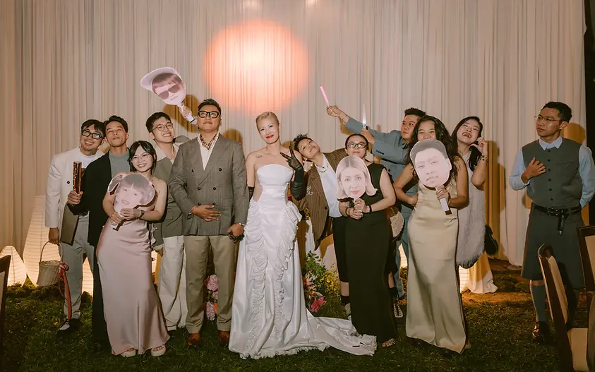 Nhóm bạn chụp ảnh cùng cặp đôi trong tiệc cưới buổi tối, nhiều người cầm bảng vui tạo không khí trẻ trung.