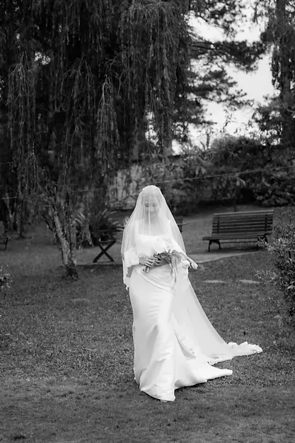 Cô dâu bước đi trong khu vườn vào lễ đường với khăn voan phủ nhẹ, khung cảnh yên bình và lãng mạn.