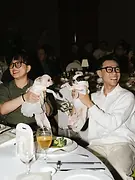 Kiss Cam 04: Two guests holding small dogs while smiling at their table during the wedding celebration.