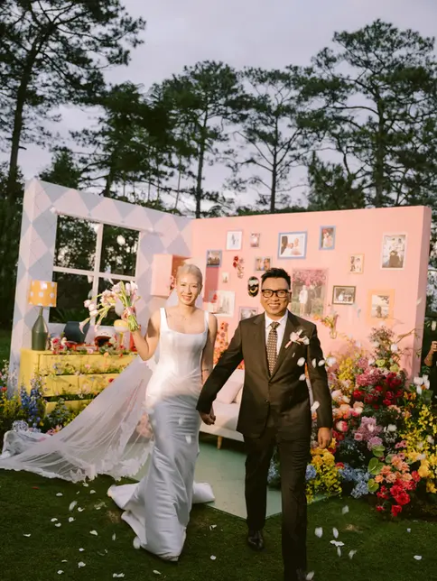 Cặp đôi nắm tay bước ra từ sân khấu cưới ngoài trời giữa cánh hoa rơi, khung hình tràn đầy năng lượng và niềm vui.