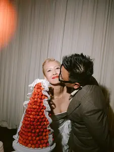 A bride smiling while the groom kisses her cheek beside a tall strawberry croquembouche at their wedding celebration.