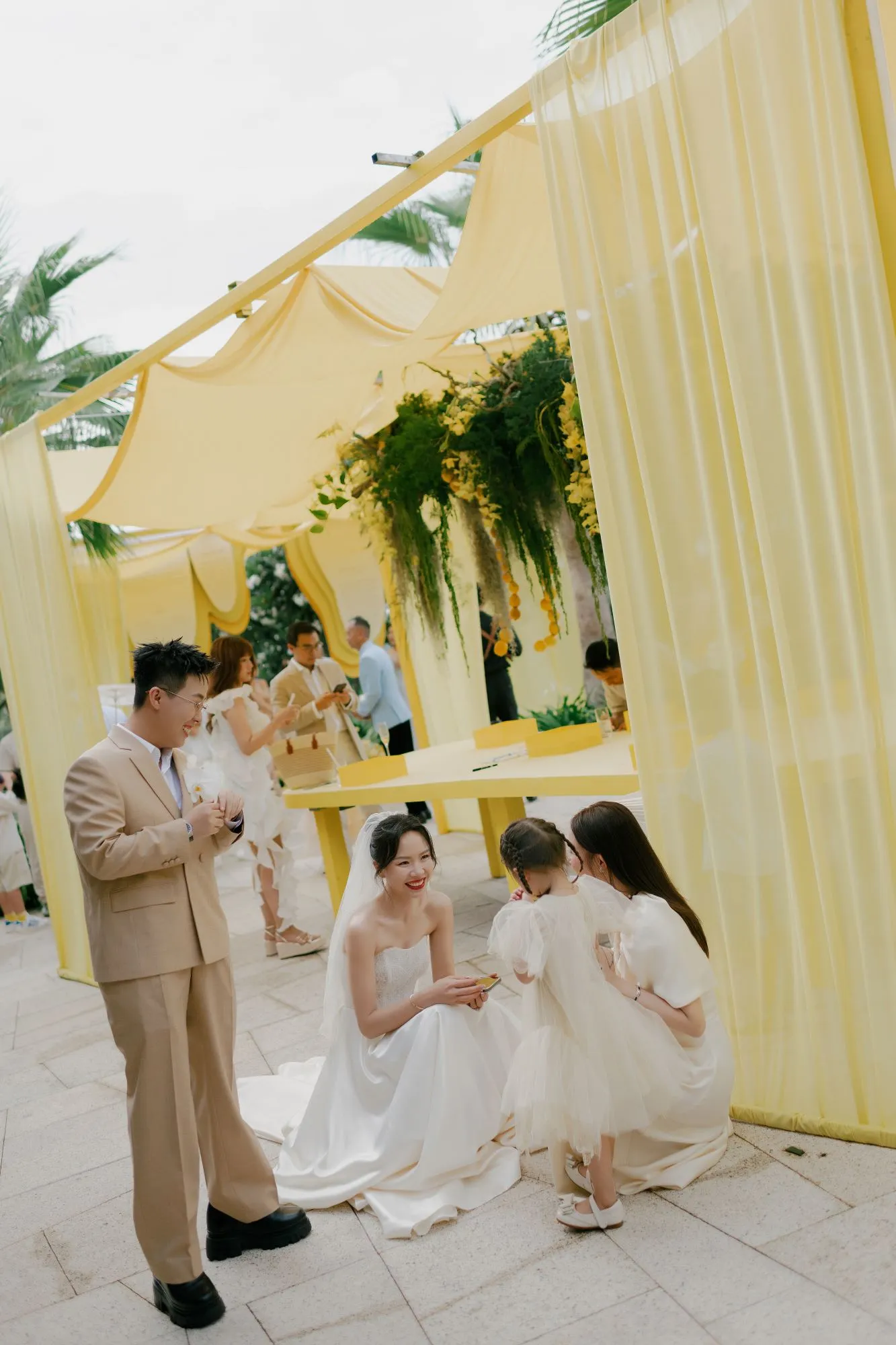 The bride kneels to greet a flower girl under soft yellow wedding décor, creating a warm and joyful pre-ceremony moment.
