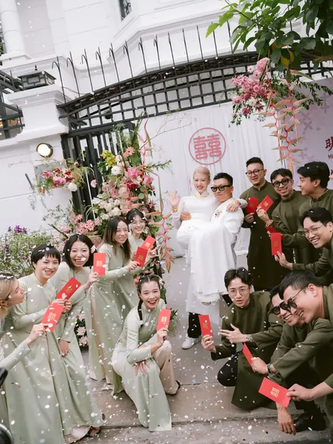 Groom lifting the bride while friends gather around laughing and holding red envelopes.