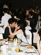 Kiss Cam 03: A couple sharing a kiss at their table while guests applaud during the wedding reception.