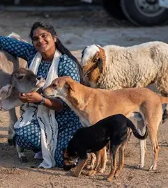 A woman smiles as dogs, goats and cows surround her. 