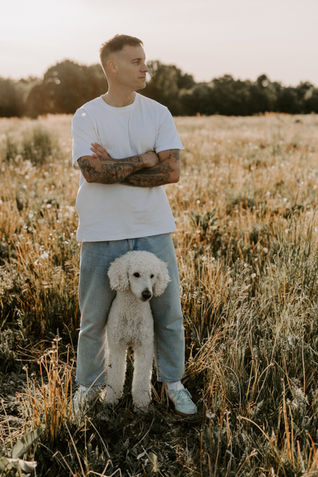 Mann mit tätowierten Armen steht im Feld, sein weißer Pudel sitzt schützend zwischen seinen Beinen – Symbol für Freundschaft, Vertrauen und Zusammenhalt.