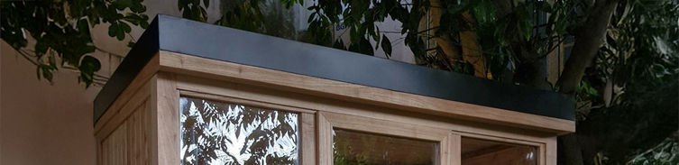 Sauna de exterior con techo plano impermeable y cubierta metálica negra, ubicado en un jardín bajo árboles.