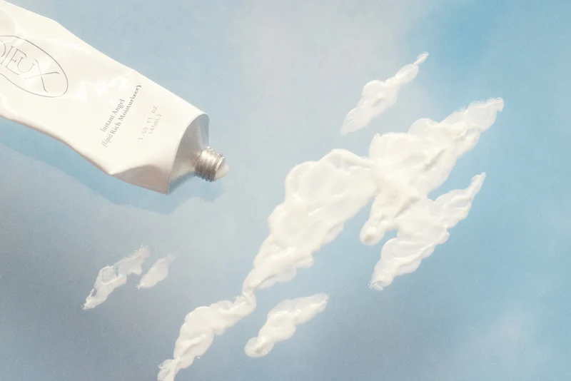 Dieux Skin moisturizer forming cloud shapes from tube, styled on surreal blue sky backdrop