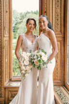 Two brides holding a white bouquet beside an ornately carved window at Manor by the Lake.