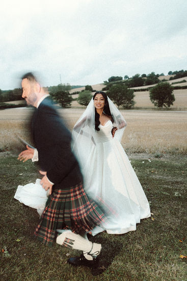 Groom in a tartan kilt moving past the smiling bride in the fields at Bury Court Barn.