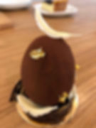 A chocolate egg dessert with gold flakes and white chocolate decoration, set on a wooden table with another dessert blurred in the background.