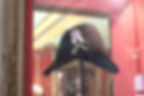 Black bicorne hat with a rosette displayed in a glass case; ornate frame, red background, museum lighting. Historical and elegant.