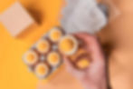 Hand holding half a peeled chocolate egg with white and yellow layers, next to a carton of similar eggs on an orange background.