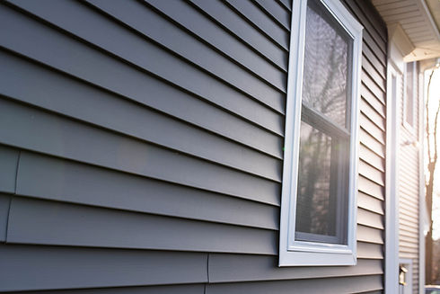 A medium shot of the facade of a house covered in gray, which highlights a white window. The scene is illuminated by the soft light of the evening, creating a warm and welcoming atmosphere