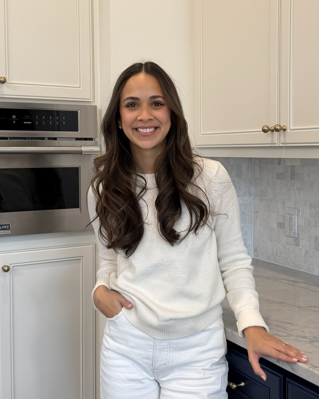 Nicole McFadden in a cream sweater, standing in a kitchen with pale cabinets and a marble counter.