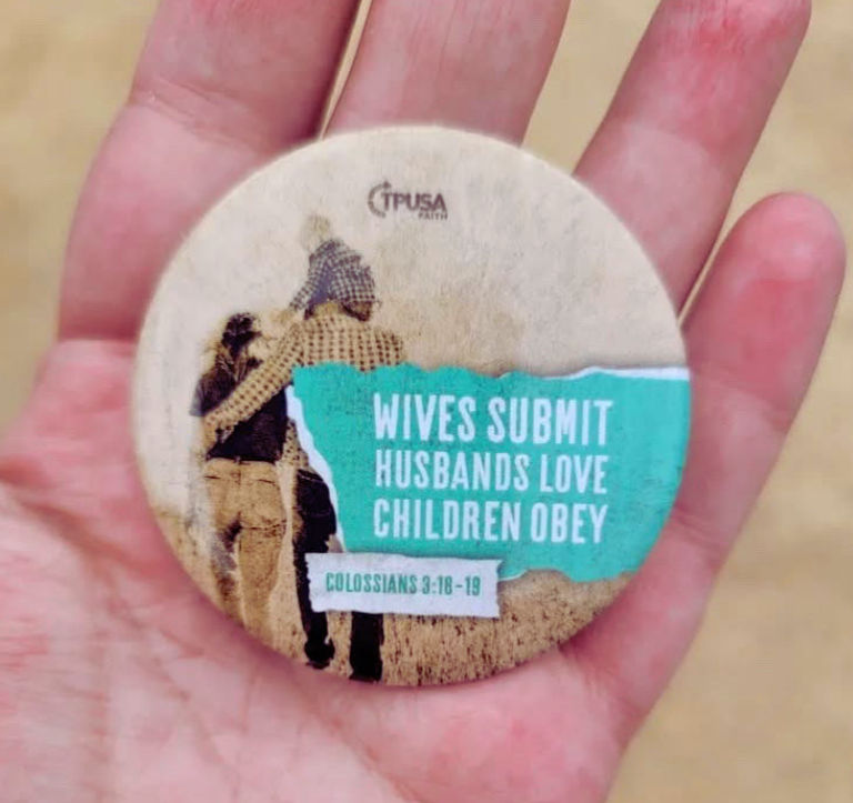 Hand holding a circular badge with an image of a couple. Text: "WIVES SUBMIT, HUSBANDS LOVE, CHILDREN OBEY, COLOSSIANS 3:18-19" on a teal background. Pin handed out from Turning Point USA.