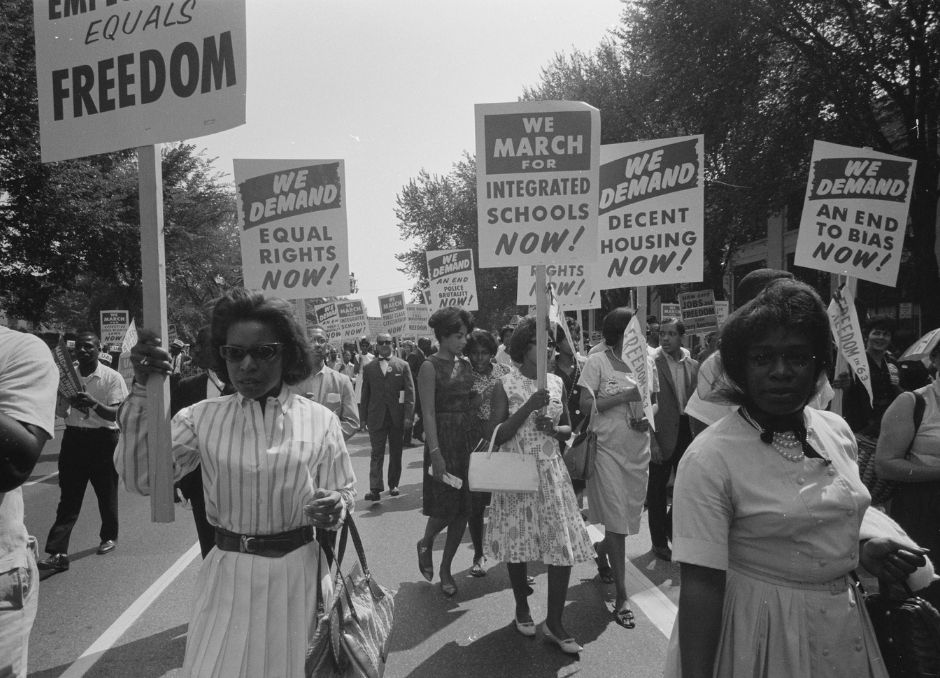 Black people marching with signs in the 1950s demanding equal rights, integrated schools, and decent housing. Black and white image, trees in the background.