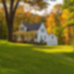 White house with porch on a green lawn, surrounded by vibrant autumn trees. Sunny day with a tranquil, peaceful atmosphere.