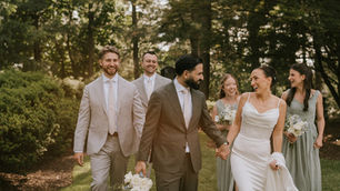 a bride and groom smile at each other as their wedding party walks behind them