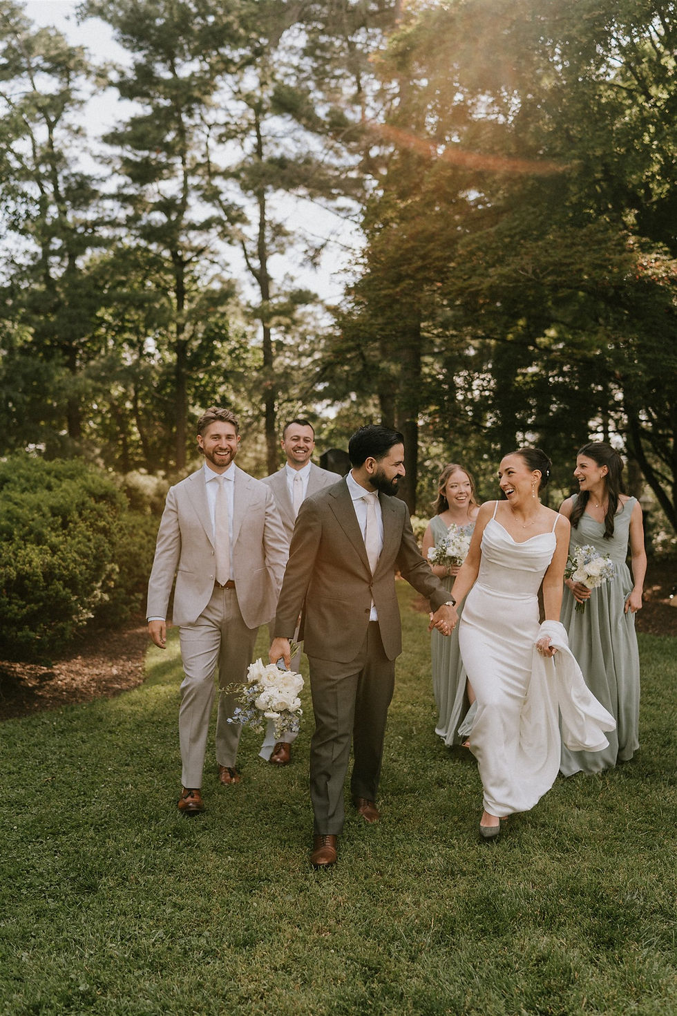 a bride and groom smile at each other as their wedding party walks behind them