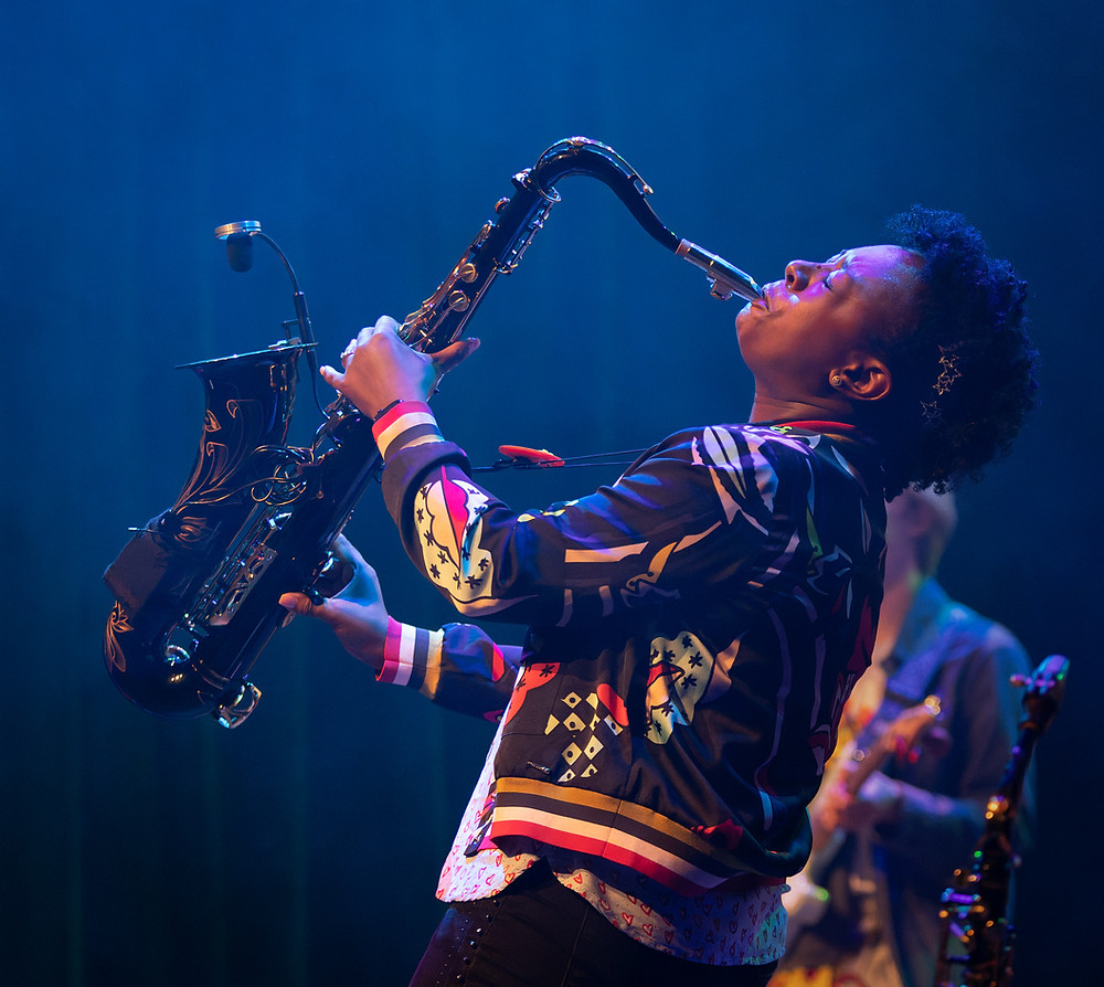 Yolanda Brown wearing a colourful jacket on stage playing Saxophone energetically.