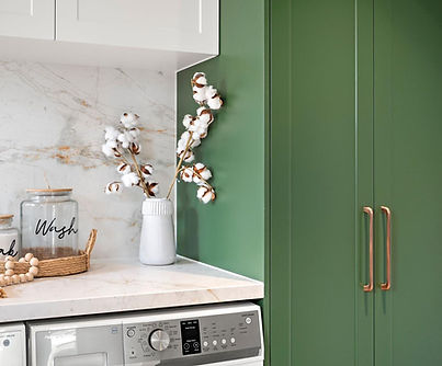 Renovated laundry room detail showcasing a sage green linen cupboard with long copper pull handles, white marble-look workbench above a front-load washing machine, and white tiled backsplash.