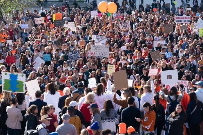 Crowded protest with diverse group holding signs against gun violence, set outdoors with trees. Orange balloons add color to the scene.