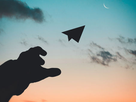 Photograph showing a silhouette of a hand launching a paper airplane against a colorful sunset sky with scattered clouds and a crescent moon. The image captures the moment of launch to demonstrate a successful product launch.